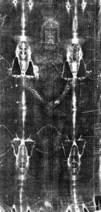 Black and white negative image of the Shroud of Turin, showing a faint full-body imprint of a man’s front and back, with arms crossed and facial features faintly visible on the fabric.