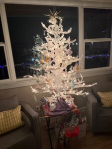 A white artificial Christmas tree decorated with lights and ornaments stands on a table between two gray armchairs, with wrapped gifts underneath and colorful string lights visible outside the window.