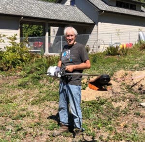 An older man stands outdoors holding a weed trimmer on a sunny day, smiling at the camera. He is surrounded by plants and dirt, with a house and a fenced yard in the background.