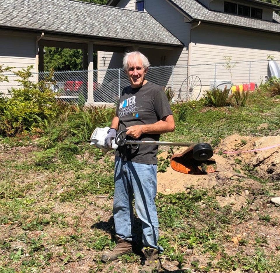 An older man stands outdoors holding a weed trimmer on a sunny day, smiling at the camera. He is surrounded by plants and dirt, with a house and a fenced yard in the background.