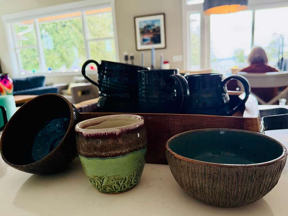 A collection of ceramic cups and bowls sits on a kitchen counter, with a window, framed photo, and a person seated in the background. Natural light fills the room.
