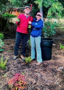 Two people stand in a garden holding gardening tools, smiling at the camera. They wear gloves, casual clothes, and hats. There are plants and a large black bin nearby, with trees and greenery in the background.
