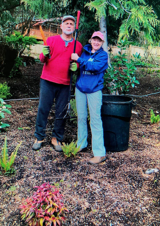 Two people stand in a garden holding gardening tools, smiling at the camera. They wear gloves, casual clothes, and hats. There are plants and a large black bin nearby, with trees and greenery in the background.