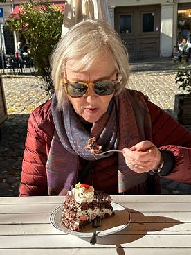 An older woman wearing sunglasses and a scarf sits at an outdoor table, about to eat a forkful of chocolate cake topped with cream and a strawberry on a sunny day.