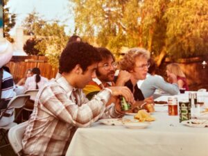 Three people sit at an outdoor table eating and drinking. They appear to be at a casual gathering or picnic. There is food, plates, and soda cans, including 7UP, on the table. Other people are in the background.
