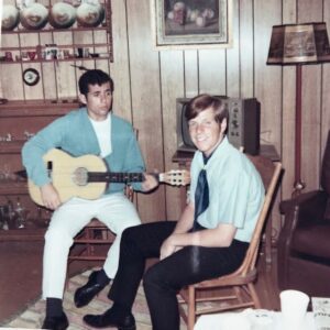 Two young men sit in a wood-paneled room; one plays a guitar while the other smiles at the camera. There is a lamp, a dresser with decorations, and an old TV in the background.