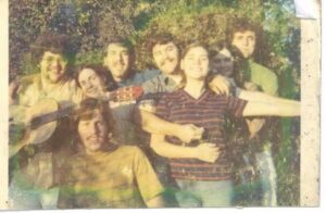 A group of seven young adults, some smiling and hugging, pose outdoors. One person is holding a guitar. The image appears faded and vintage, with greenery in the background.